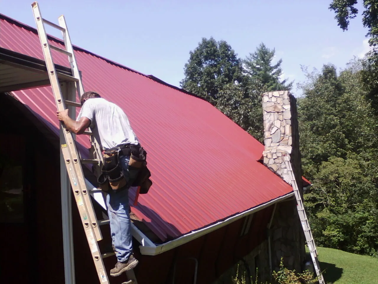 Roofer on a ladder beside a red metal roof and stone chimney — Soffit & Fascia Repair by Roofing Brothers in Niles